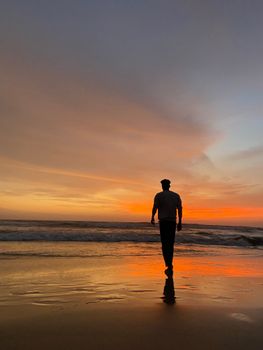 A contemplative man walks along a sandy beach at sunset, silhouetted against the colorful sky.