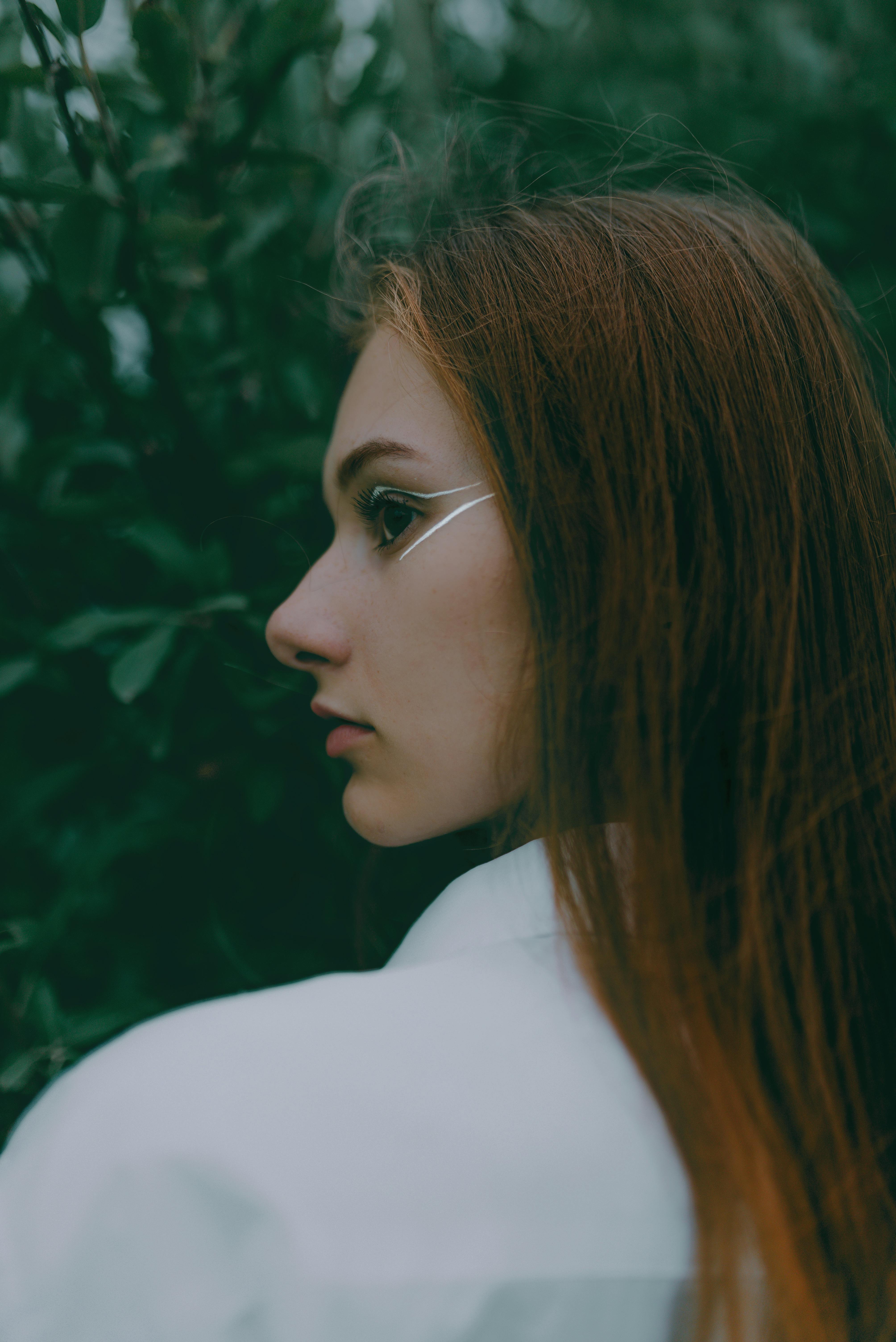 A captivating side profile of a young woman with makeup, surrounded by lush greenery.