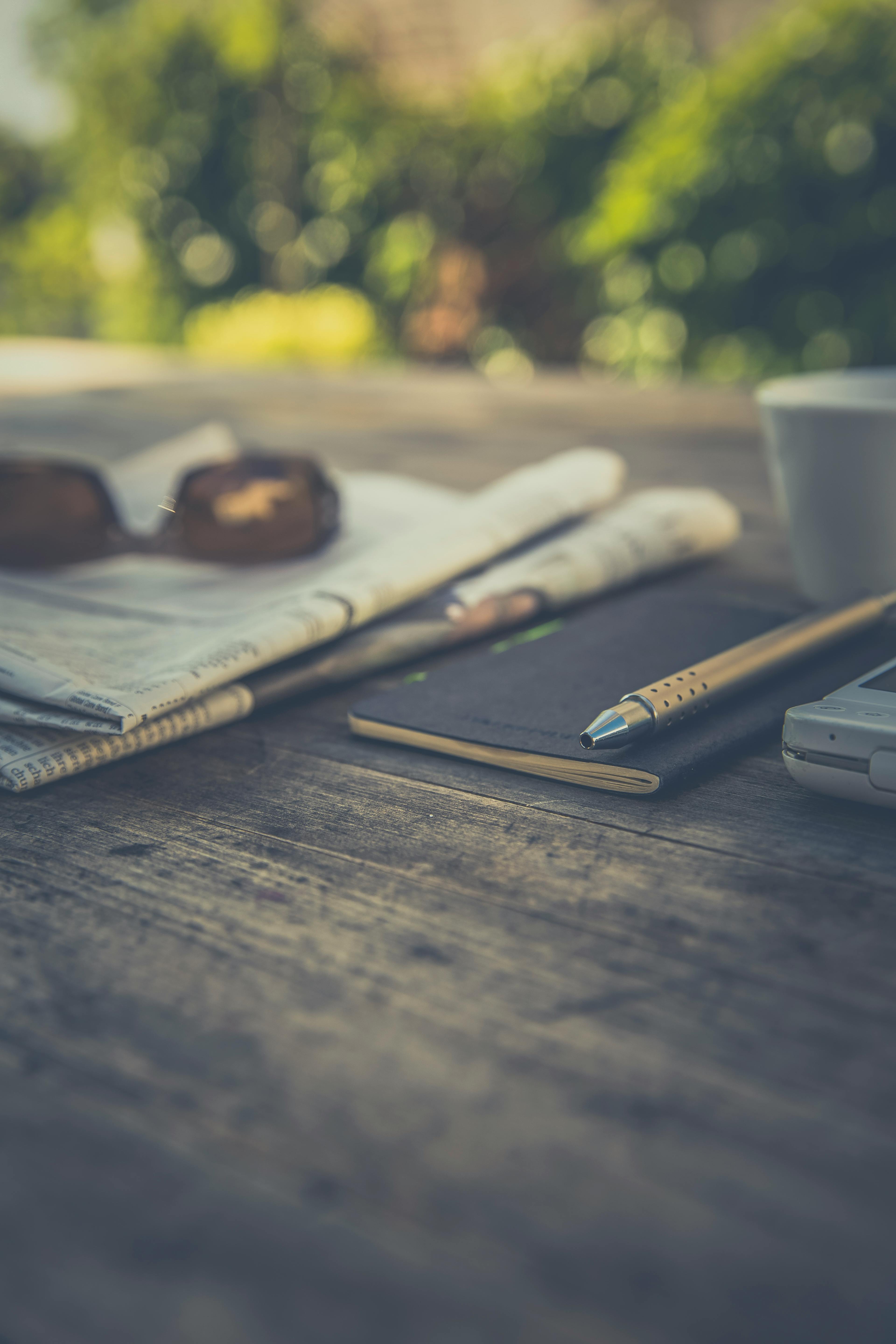 木桌上摆放着报纸、笔、智能手机和咖啡杯，环境宁静自然