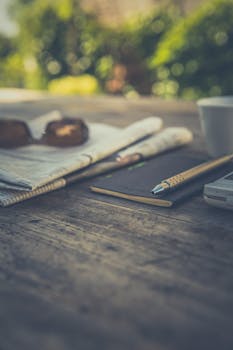 木桌上摆放着报纸、笔、智能手机和咖啡杯，环境宁静自然