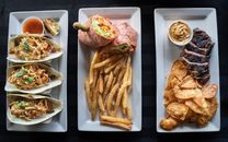 A top view of diverse dishes including tacos, a wrap with fries, and steak with chips on white plates.