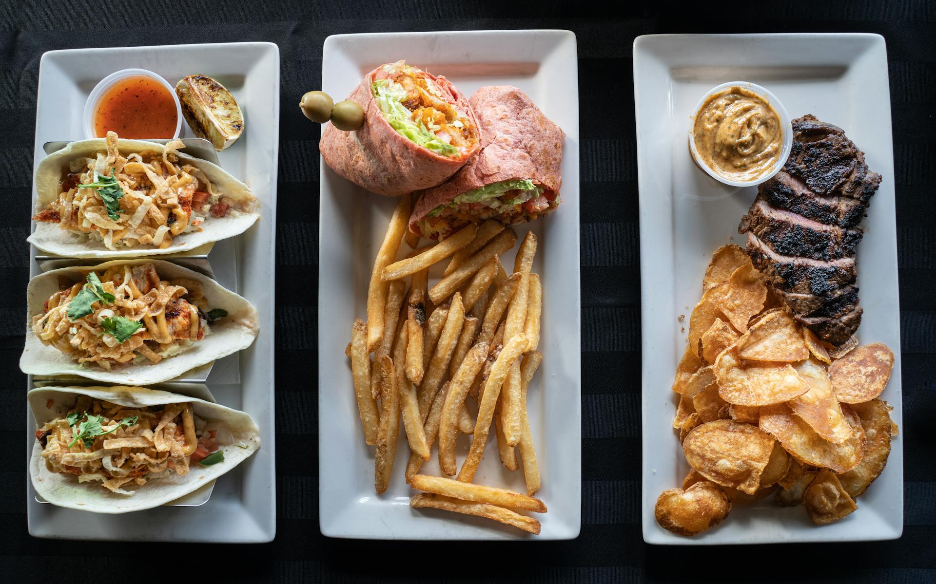 A top view of diverse dishes including tacos, a wrap with fries, and steak with chips on white plates.