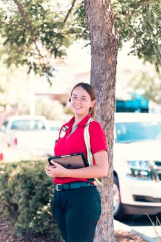 Young woman in a red polo with earphones holding a tablet outdoors near cars and a tree.