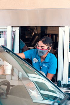 A fast-food drive-thru employee wearing a mask serves a customer. COVID-19 safety measures in place.