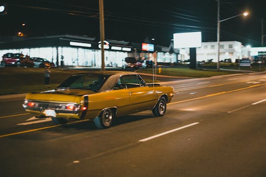 A classic vintage coupe car driving down a well-lit city street at night with blurred background lights.