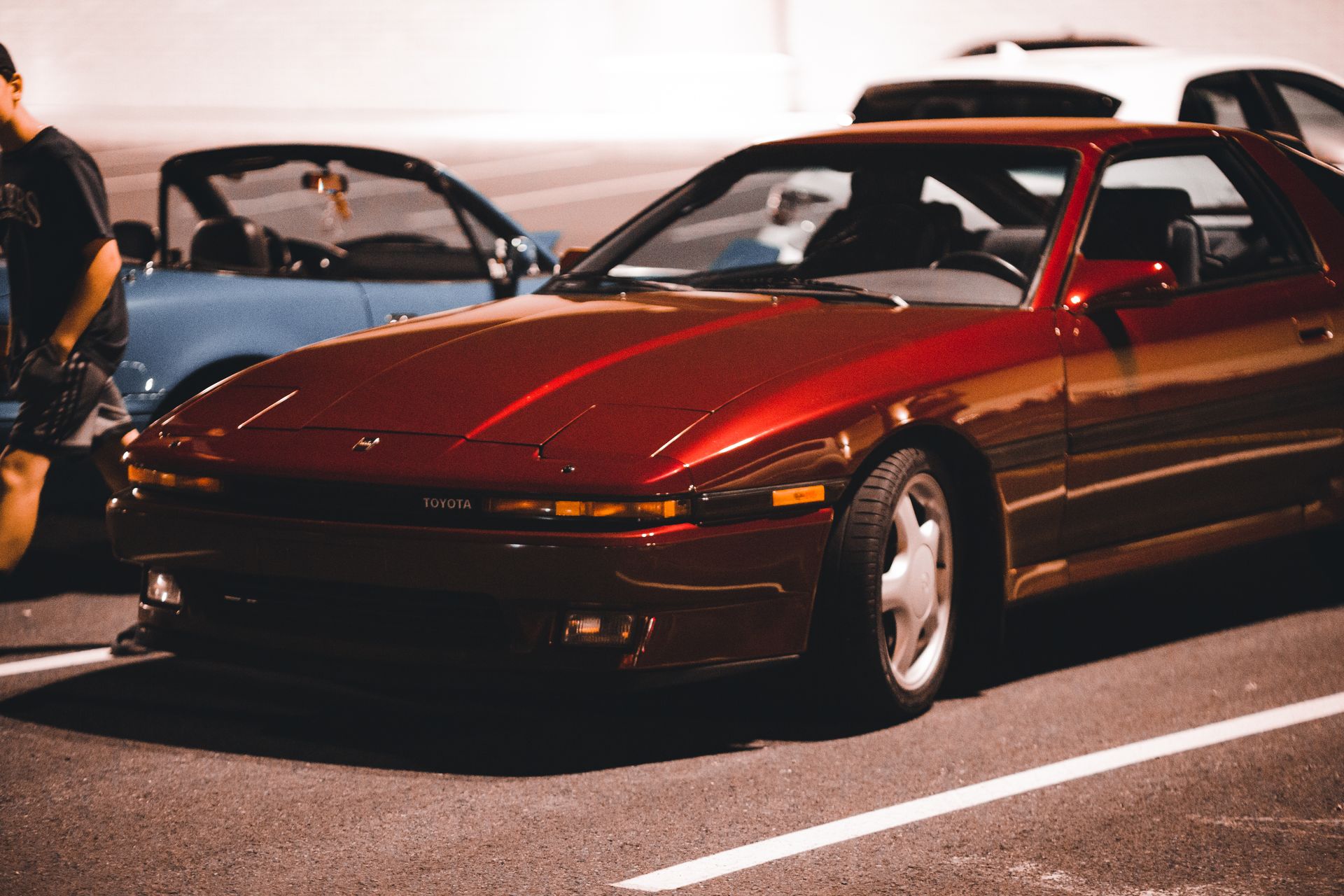 Classic red Toyota car parked in a lot at night with a person nearby.