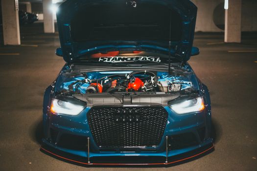 Blue sports car with open hood displaying engine in dimly lit urban setting.