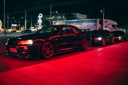 A lineup of modified sport cars parked under striking neon lights at night, showcasing automotive elegance.
