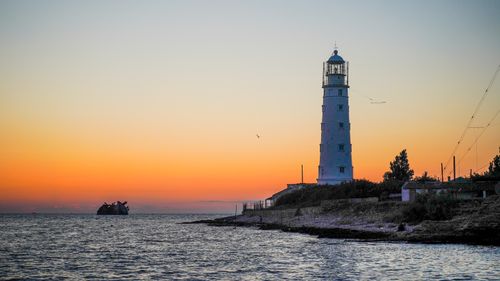 Free A picturesque lighthouse silhouetted against a vibrant sunset sky by the sea. Stock Photo