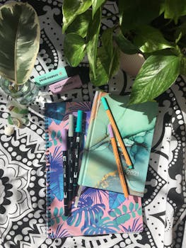 Colorful journals and pens arranged creatively on patterned fabric with lush green plants.