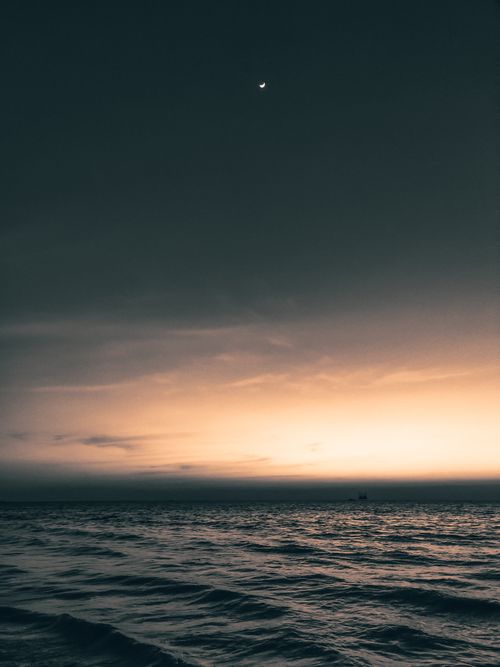 Free A tranquil ocean scene at twilight featuring a crescent moon and gentle waves under a darkening sky. Stock Photo