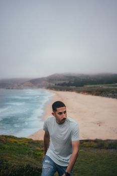 Young man with a mustache walks near the foggy beach cliffs, wearing ripped jeans and a gray shirt.