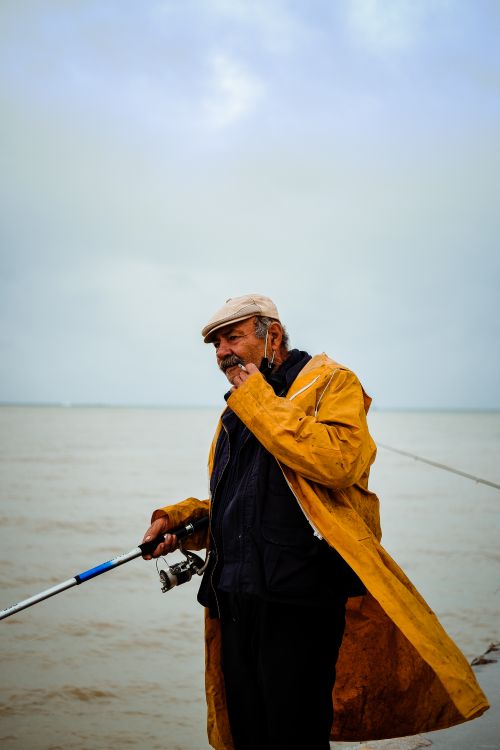 Free Side view of old male fisher in yellow raincoat and mask standing near rippling water with fishing rod and catching fish in daylight Stock Photo