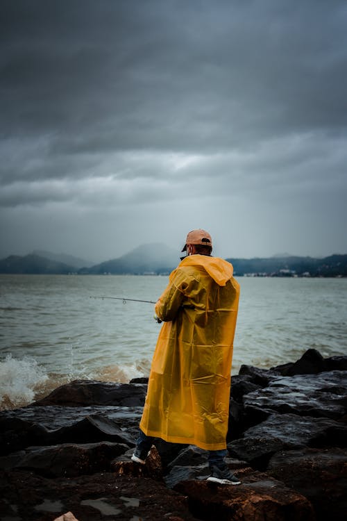 Back view of unrecognizable male in yellow raincoat standing on rocky coast against waving sea and holding fishing rod while fishing in stormy weather