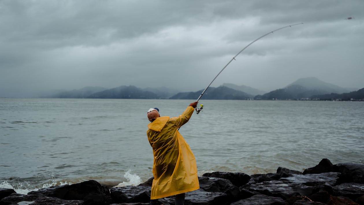Back view of unrecognizable fisherman in raincoat standing on rocky coast near waving sea and catching fish with fishing rod in stormy weather