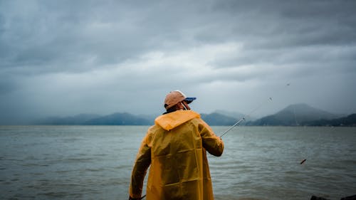 Back view of unrecognizable fisherman in yellow raincoat and mask casting fishing rod on rippled water against view of misty mountains and cloudy sky