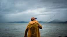 Back view of unrecognizable fisherman in yellow raincoat and mask casting fishing rod on rippled water against view of misty mountains and cloudy sky