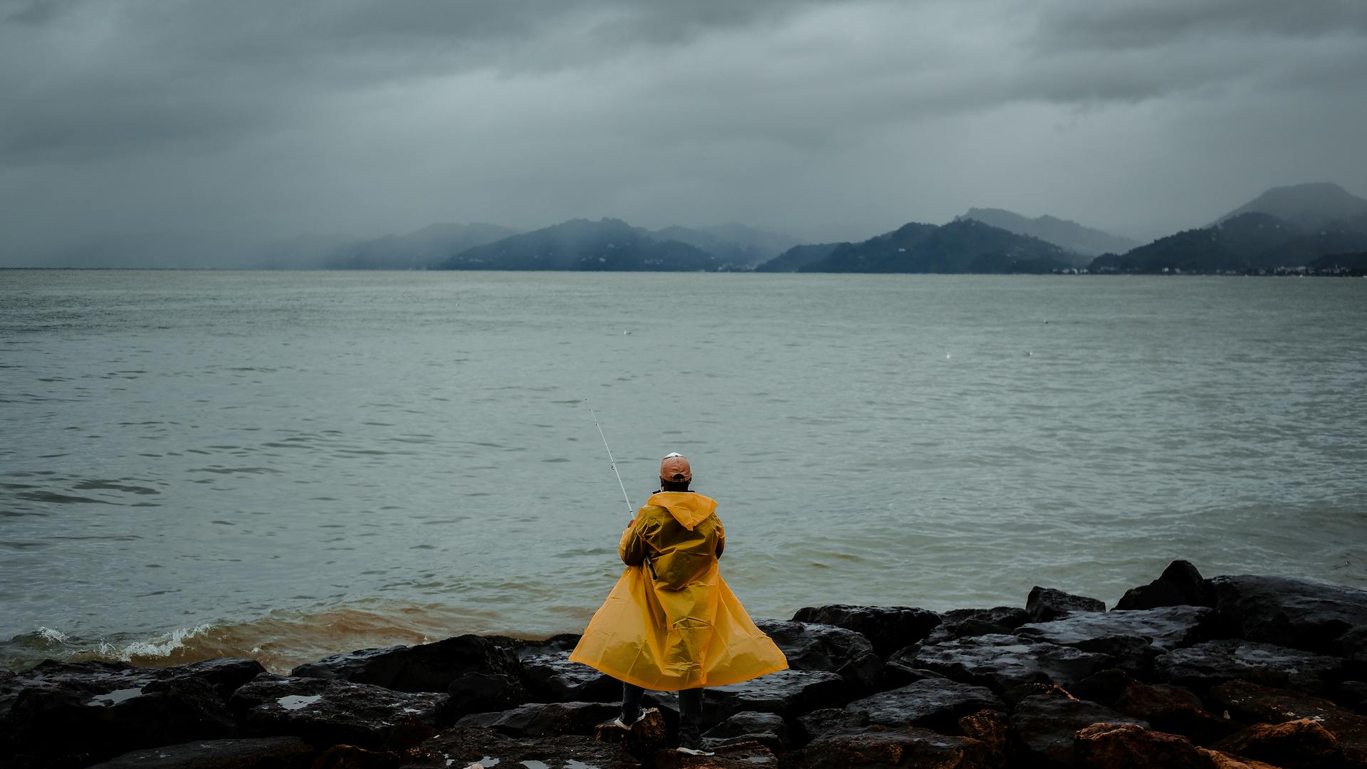 Back view of anonymous male fisher in yellow raincoat standing on rocky coast of sea and fishing in windy cloudy day