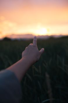 A hand pointing towards a vibrant sunset over a grassy field, creating a serene atmosphere.