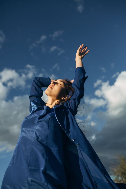 Free A woman in a blue dress enjoys the outdoors with arms raised under a clear sky. Stock Photo