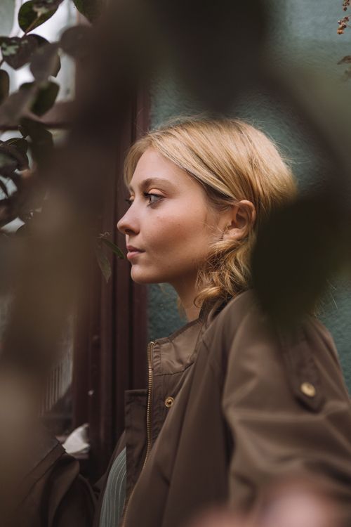 Free A young woman in a brown jacket thoughtfully gazes into the distance, surrounded by greenery. Stock Photo