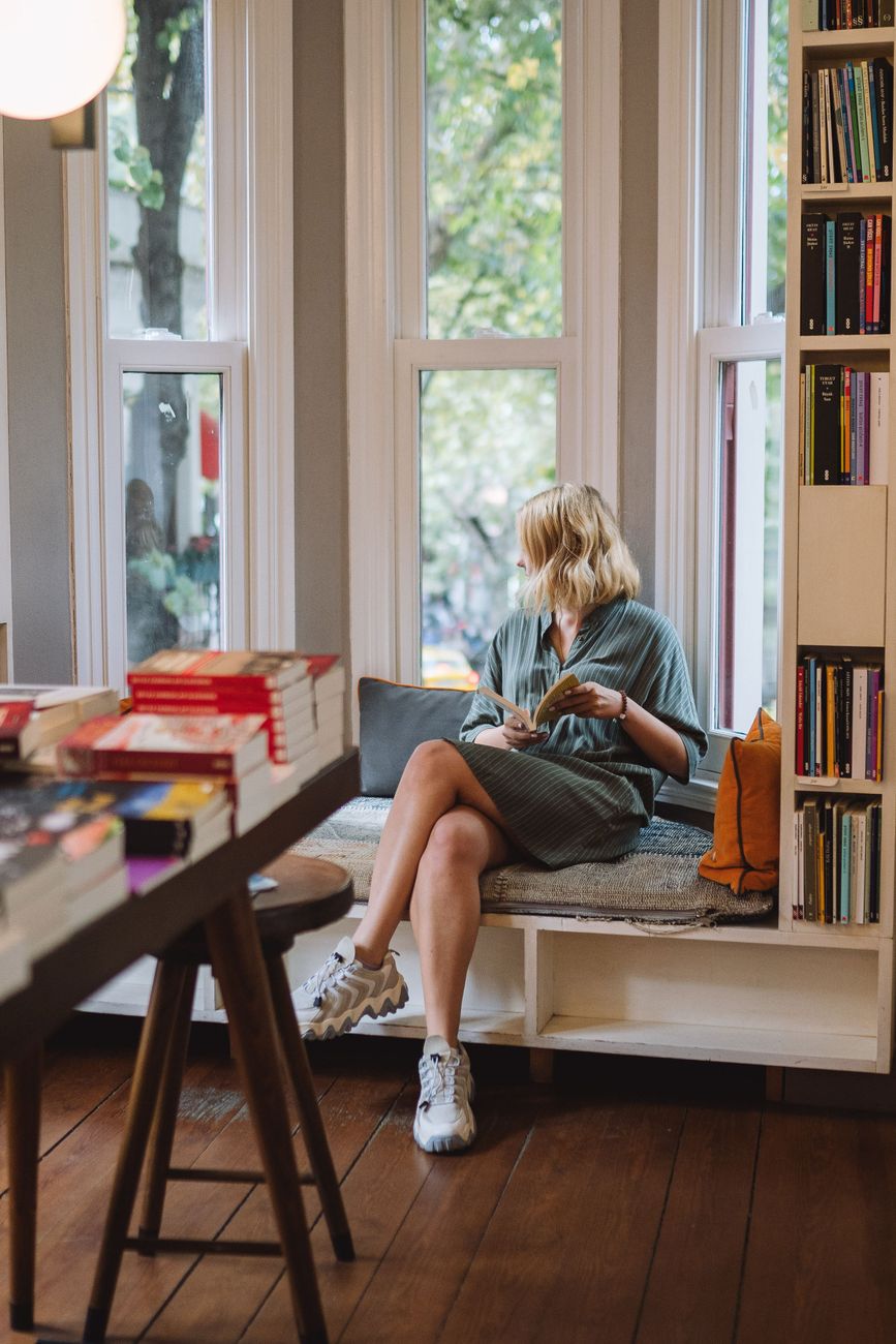 A woman enjoys reading in a cozy bookstore window nook, surrounded by books.