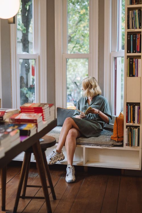 Free A woman enjoys reading in a cozy bookstore window nook, surrounded by books. Stock Photo