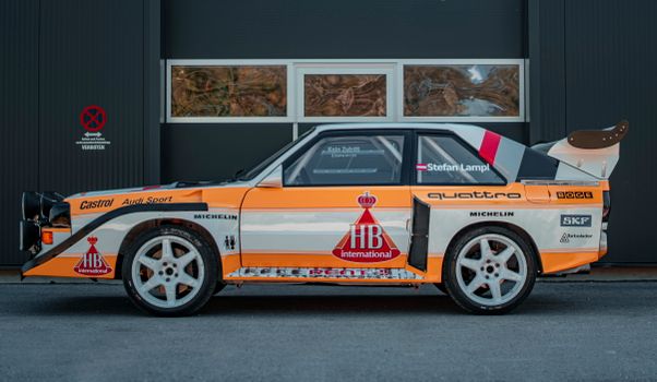 Orange and white Audi Sport Quattro S1 rally car parked outdoors with logos and decals.