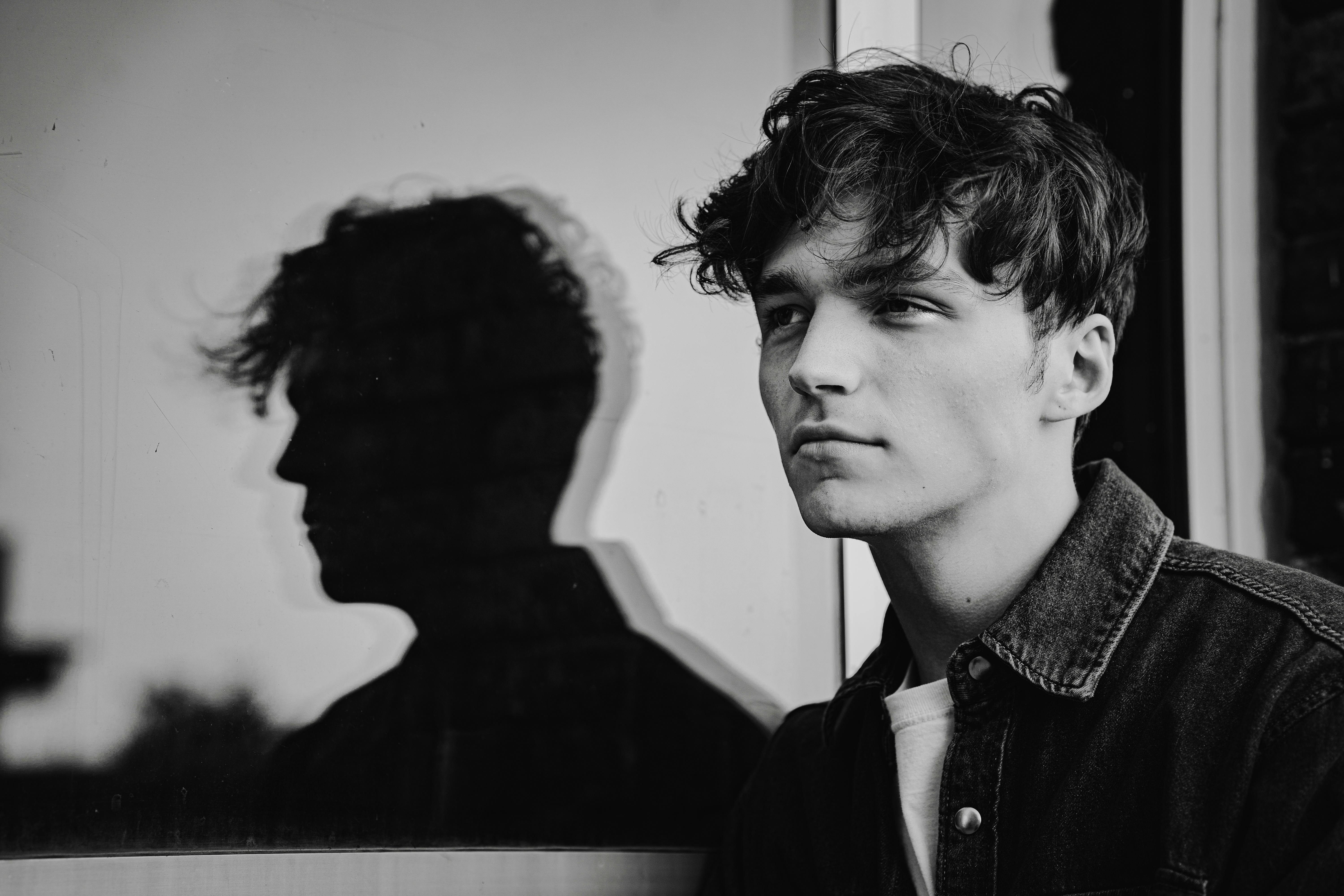 Black and white portrait of a young man with his shadow reflected on a window, evoking introspection.