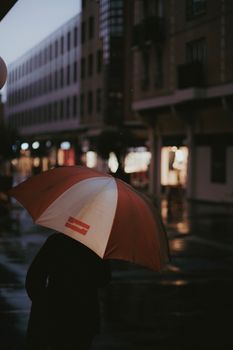 A solitary figure holding an umbrella walks through a city street at dusk, reflecting a moody, rainy atmosphere.