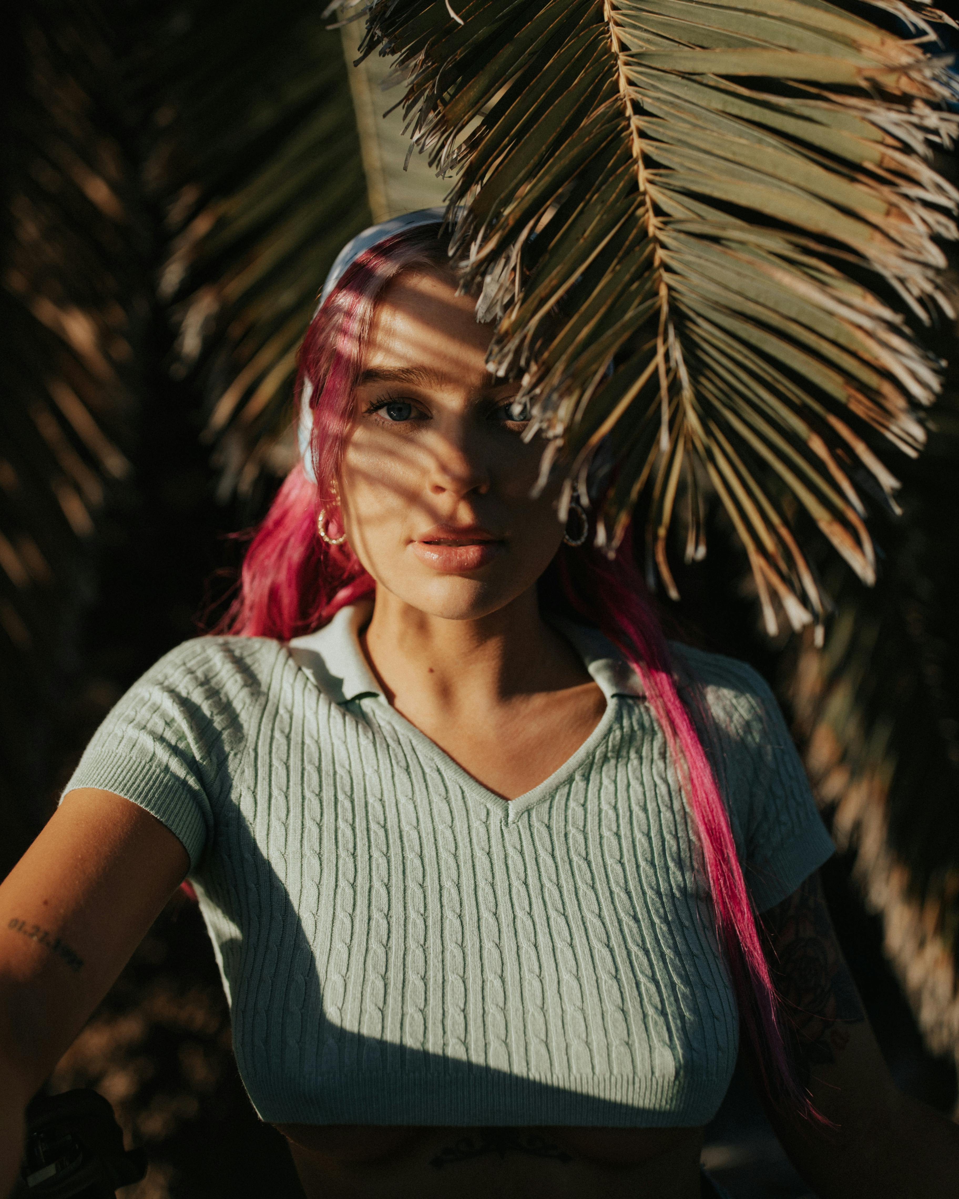 Stylish woman with pink hair poses in sunlight under palm leaves.