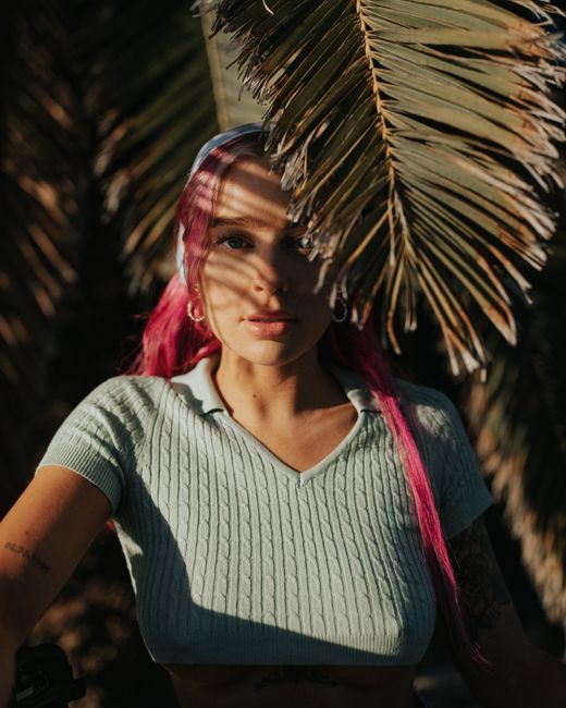 Stylish woman with pink hair poses in sunlight under palm leaves.