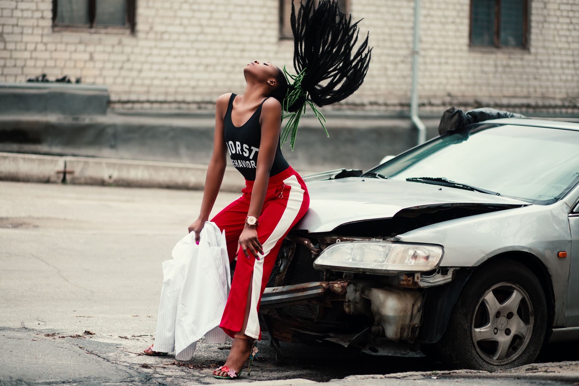 Fashionable woman with braided hair poses dynamically by a damaged car in an urban setting.