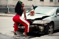 一位穿着红裤子的非裔美国妇女自信地在一辆损坏的汽车旁摆姿势，背景是城市环境