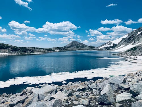 Gray Rocky Shore Across Blue Lake Under Blue and White Cloudy Sky