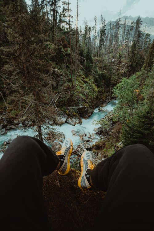 Free Point of view shot of a person sitting above a forest river with visible shoes and lush surroundings. Stock Photo