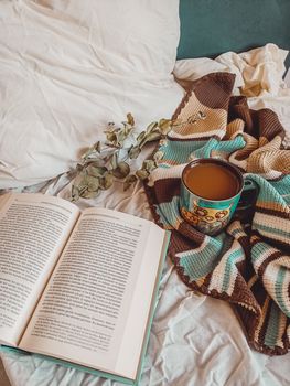 A cozy setup with an open book, warm drink, and blanket on a bed, perfect for reading and relaxing.