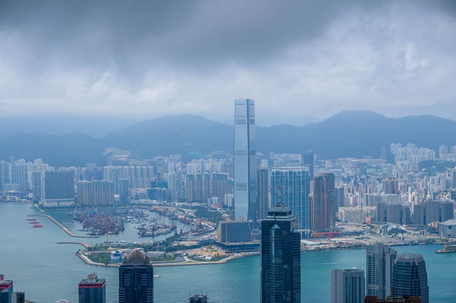 Aerial view of the iconic Hong Kong skyline and Victoria Harbor under overcast skies.