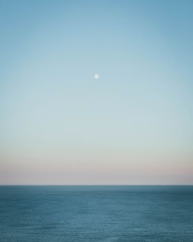 A calm ocean view under a clear blue sky during sunset with a visible full moon.