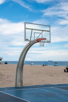 A basketball hoop stands on a sandy beach with a clear ocean view, perfect for a summer day game.