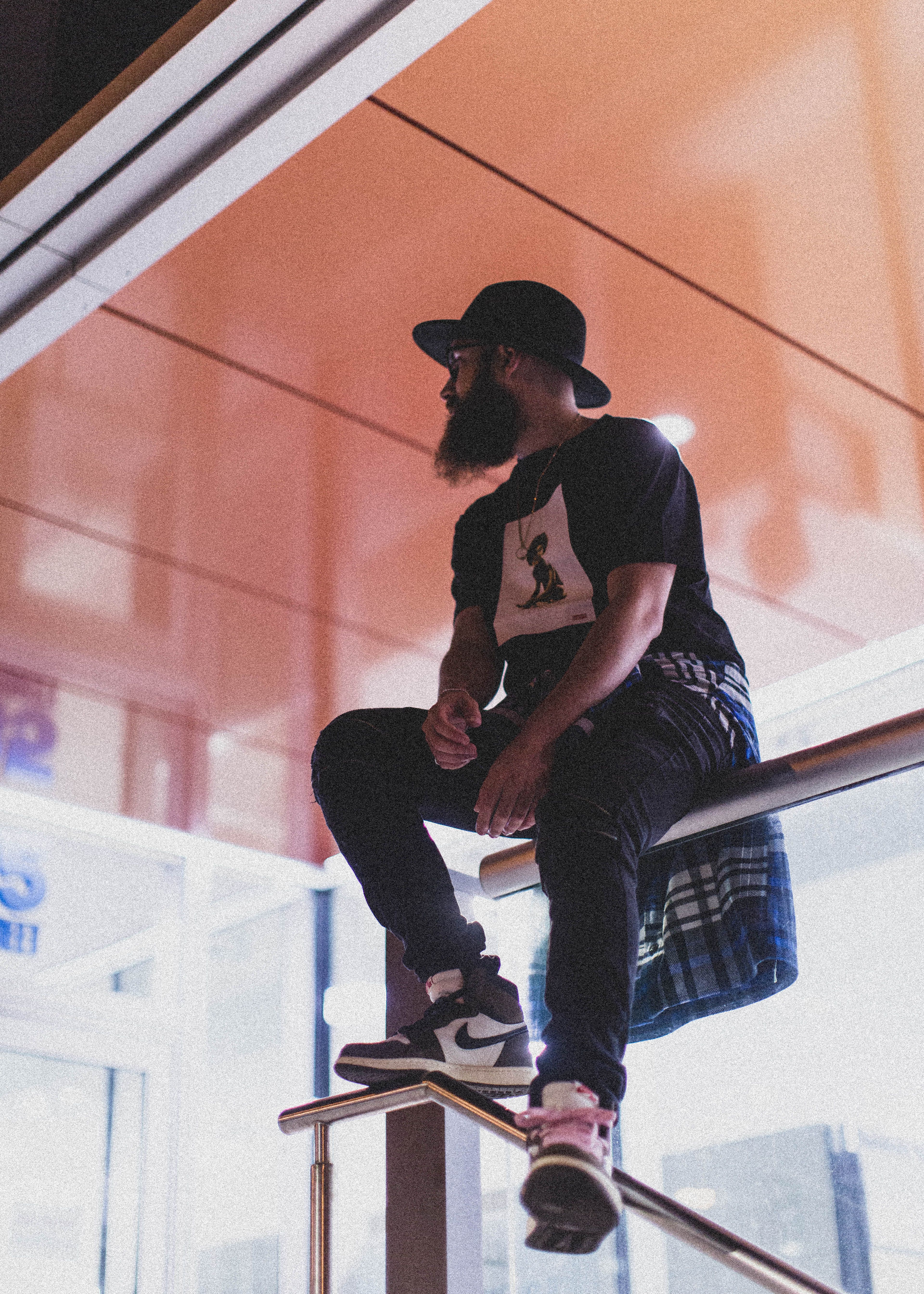 Bearded man in streetwear and hat sitting on a handrail indoors.