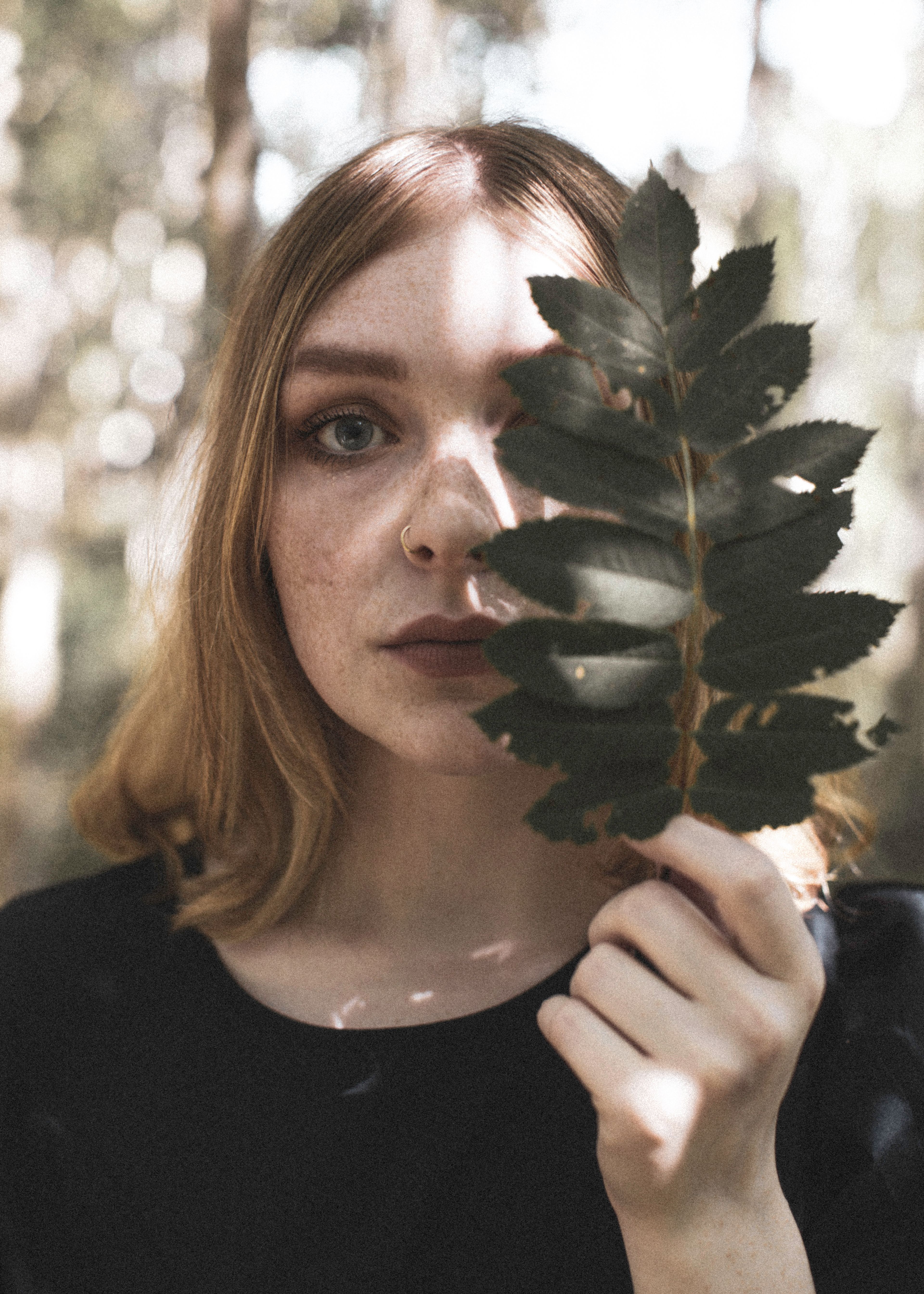 A young woman stands in a forest holding a leaf, sunlight casting shadows across her face.
