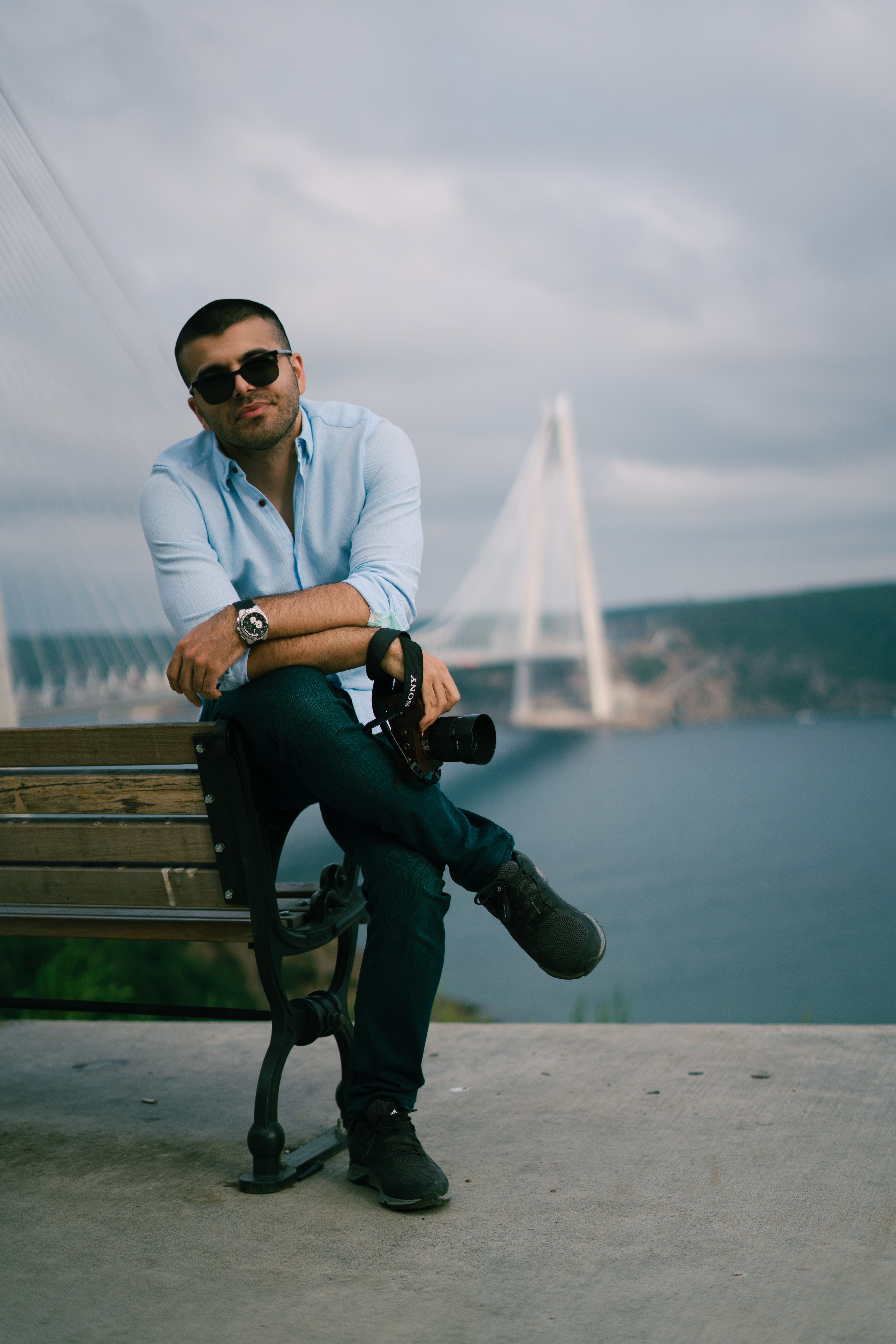 Man in sunglasses relaxes on bench with bridge and ocean backdrop, holding a DSLR camera