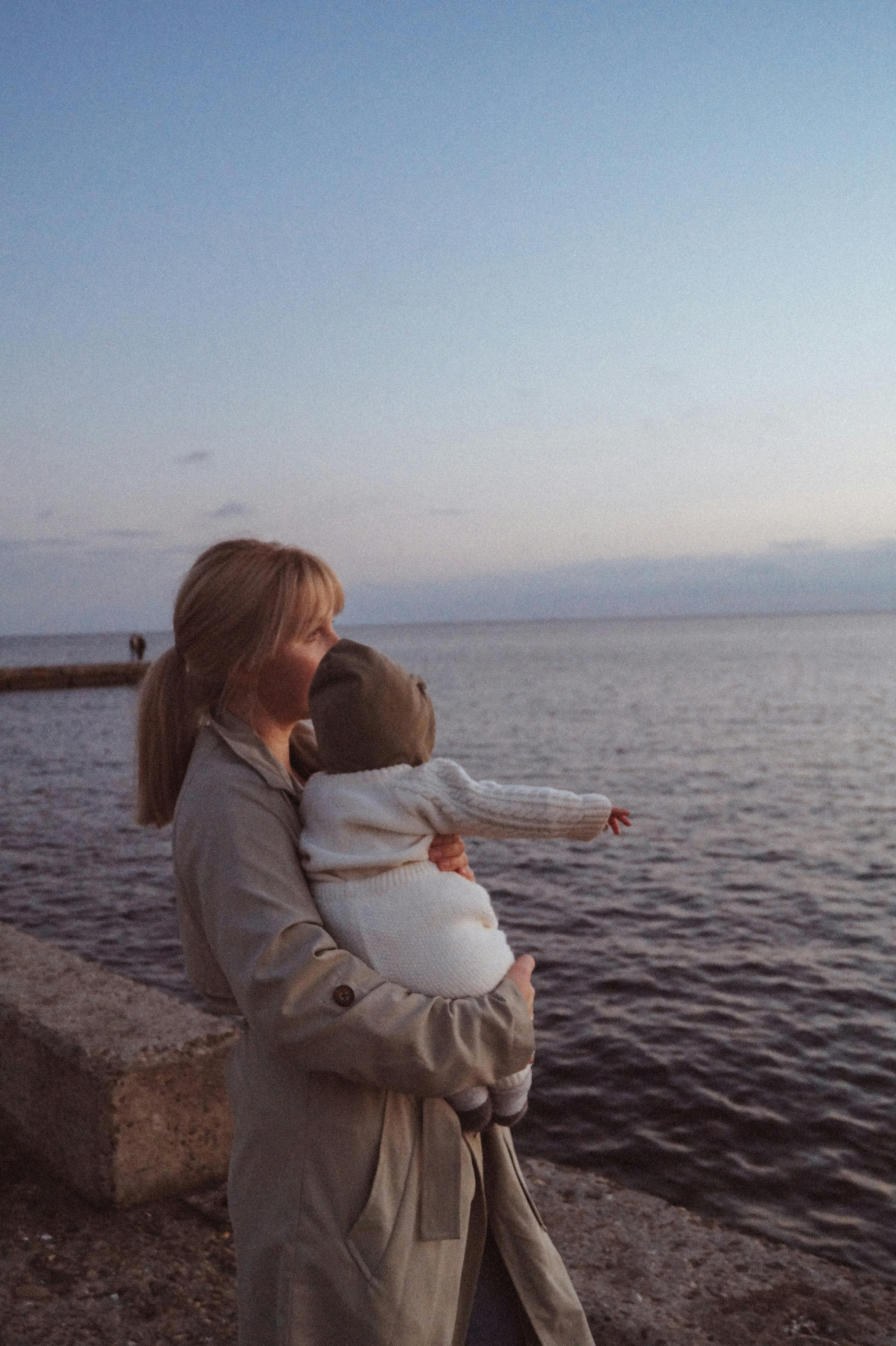 A mother holds her baby near the ocean during a calm and serene sunset.
