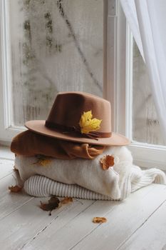 Brown hat and cozy sweaters stacked indoors by a window, decorated with autumn leaves.