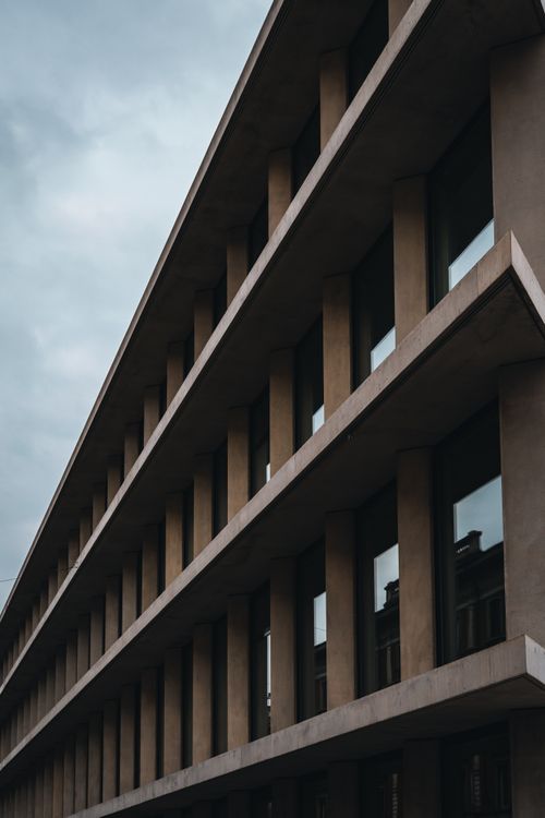 Free A modern building facade showcasing concrete and glass with reflective elements under a cloudy sky. Stock Photo