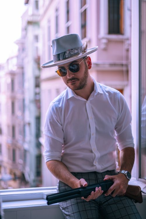 Free A fashionable man wearing a hat and sunglasses, holding a shotgun by a window in an urban environment. Stock Photo