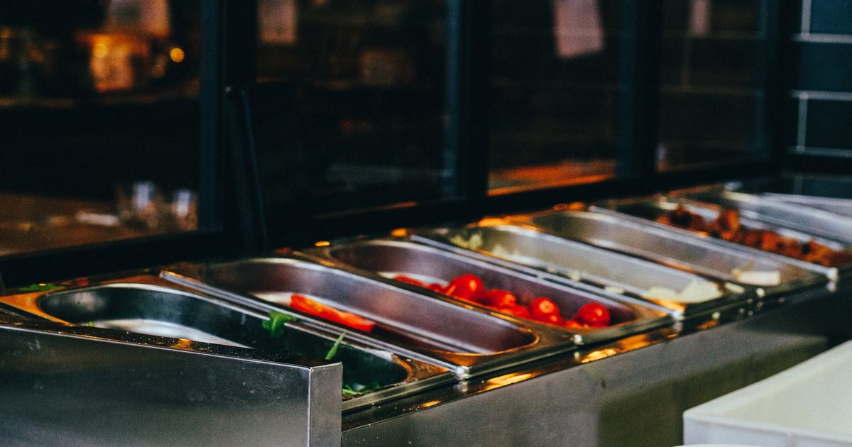 Balcão de buffet de aço inoxidável fotografado de lado, em luz baixa. As cubas metálicas estão quase vazias: uma com folhas verdes e uma fatia de tomate, outra com alguns tomates-cereja, a do fundo com restos de carne. O vidro de proteção e o salão escuro do restaurante aparecem desfocados atrás