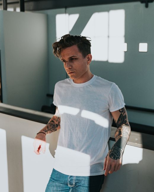 Young man with tattoos wearing white shirt in modern interior, illuminated by natural light.
