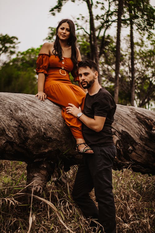 Man Standing Next to Woman in Orange Dress Sitting on a Log